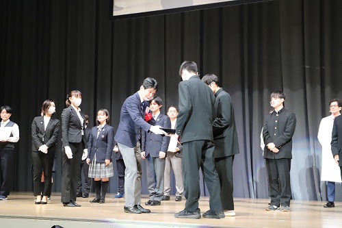 写真：大賞を受賞した伊勢崎商業高校の並里さん、向山さんに賞状及びトロフィーを贈呈する山本知事